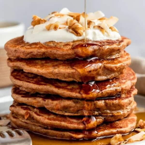 A stack of pancakes topped with whipped cream, chopped nuts, and syrup on a plate with a fork beside it; a cup is in the background.