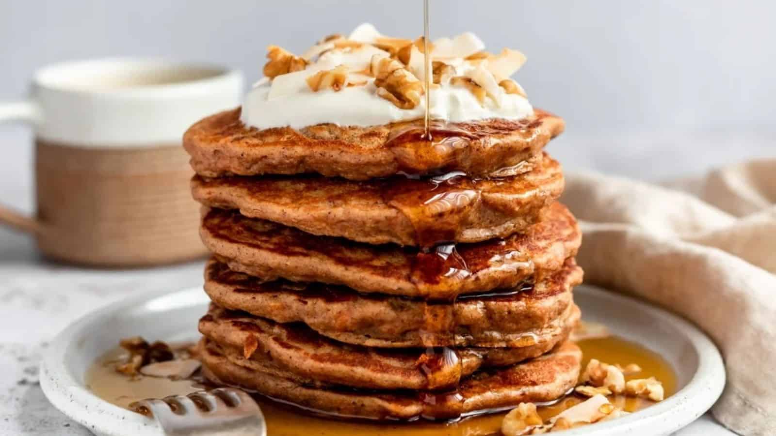 A stack of pancakes topped with whipped cream, chopped nuts, and syrup on a plate with a fork beside it; a cup is in the background.