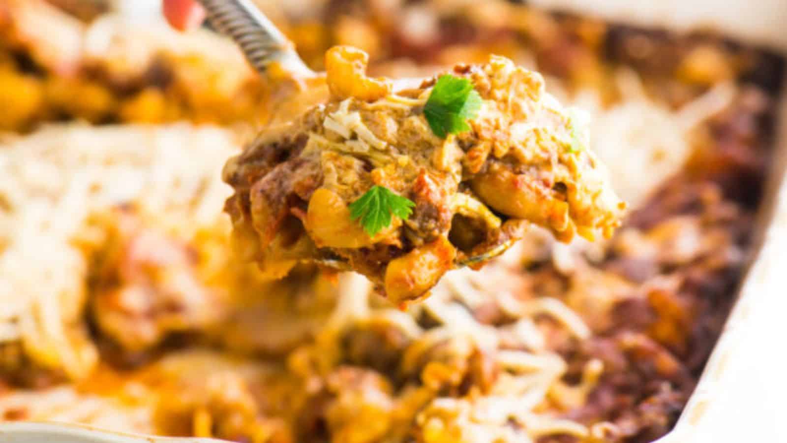 A serving spoon lifting a portion of baked pasta casserole with melted cheese, herbs, and pieces of meat over a white baking dish.