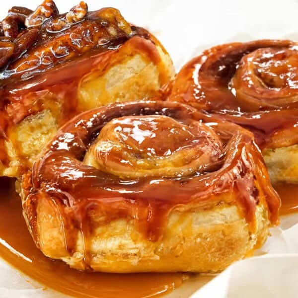 Three glazed caramel cinnamon rolls drizzled with sticky caramel and topped with pecans, resting on white parchment paper.
