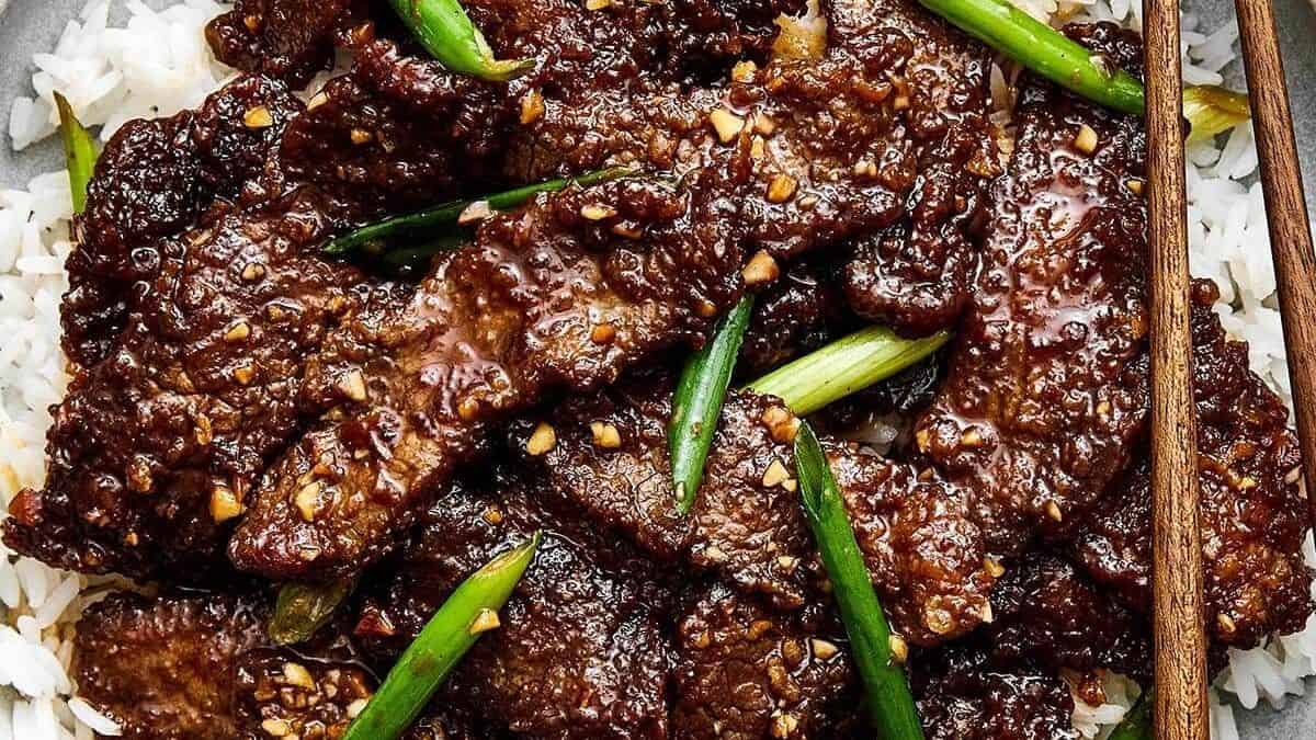 Slices of cooked beef with sauce, chopped garlic, and green onions served over white rice, accompanied by wooden chopsticks.