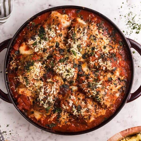 A round casserole dish filled with baked chicken parmesan, topped with tomato sauce, melted cheese, and herbs sits on a marble countertop.