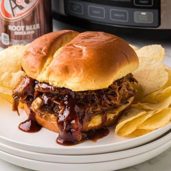 A pulled pork sandwich topped with barbecue sauce sits on a white plate beside a pile of potato chips. In the background, there is a slow cooker, a barbecue sauce bottle, and a can of root beer.