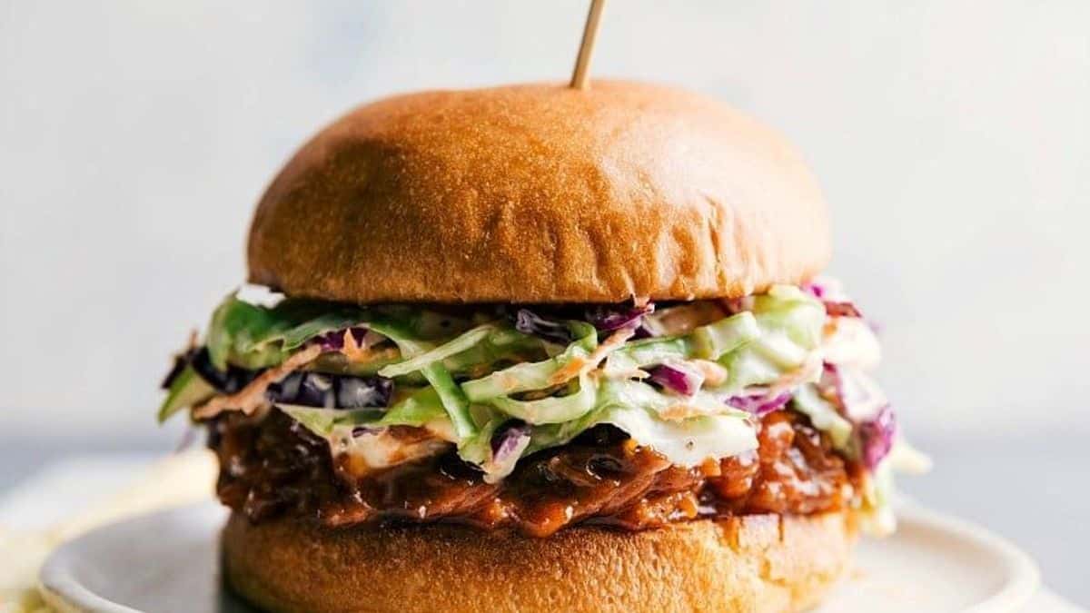 A pulled pork sandwich topped with coleslaw sits on a white plate and is held together with a wooden skewer.