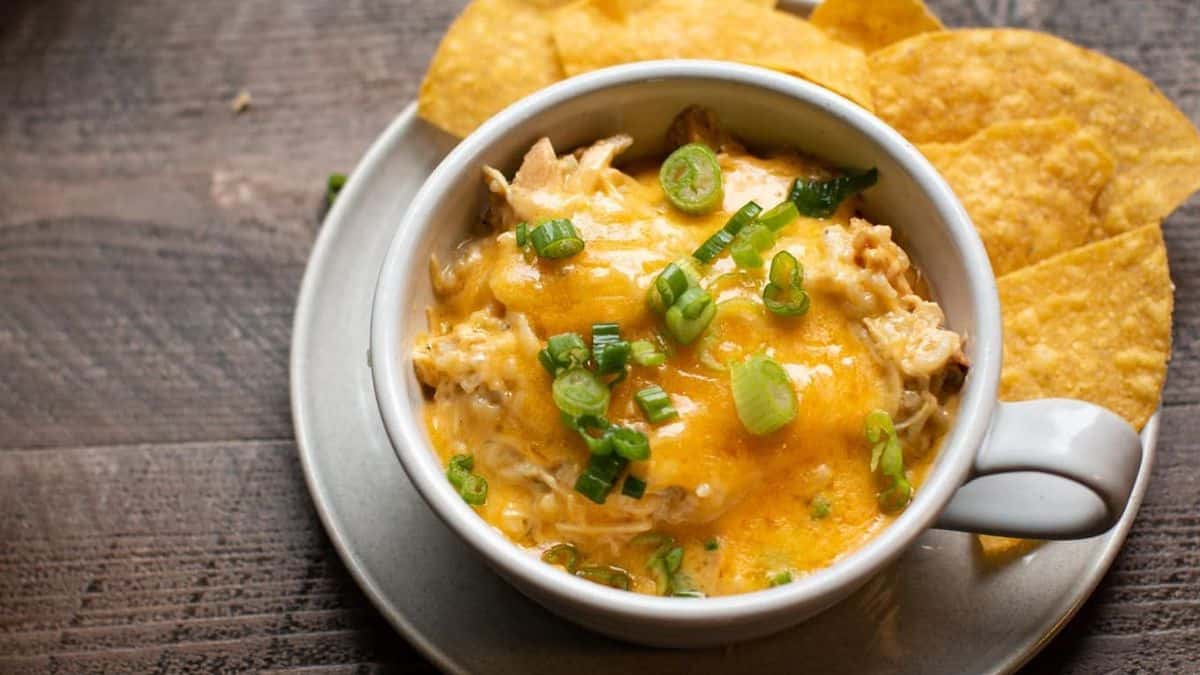 A cup filled with creamy dip topped with chopped green onions, surrounded by tortilla chips on a plate.