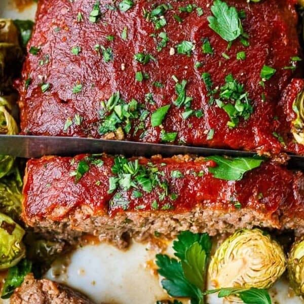 A glazed ground beef meatloaf garnished with parsley is being sliced with a knife. Roasted Brussels sprouts surround the meatloaf in a white dish.