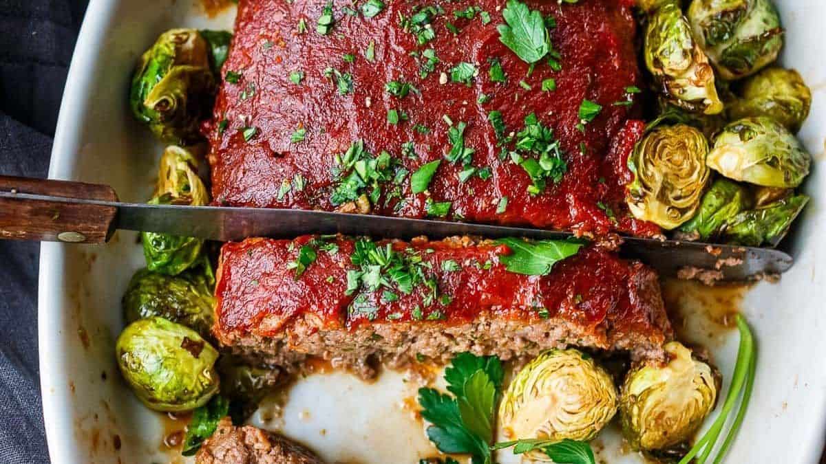 A glazed ground beef meatloaf garnished with parsley is being sliced with a knife. Roasted Brussels sprouts surround the meatloaf in a white dish.