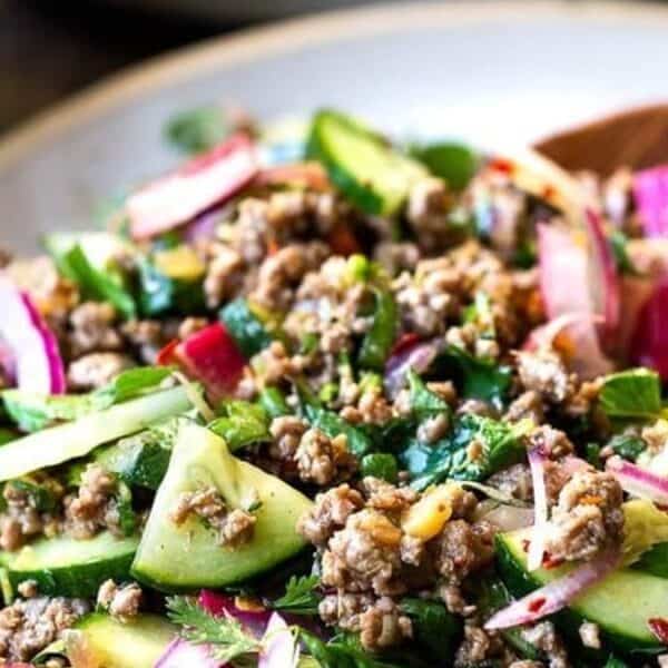 A bowl of ground meat salad mixed with cucumber slices, red onion, fresh herbs, and watermelon radish, accompanied by a wooden spoon on the side.