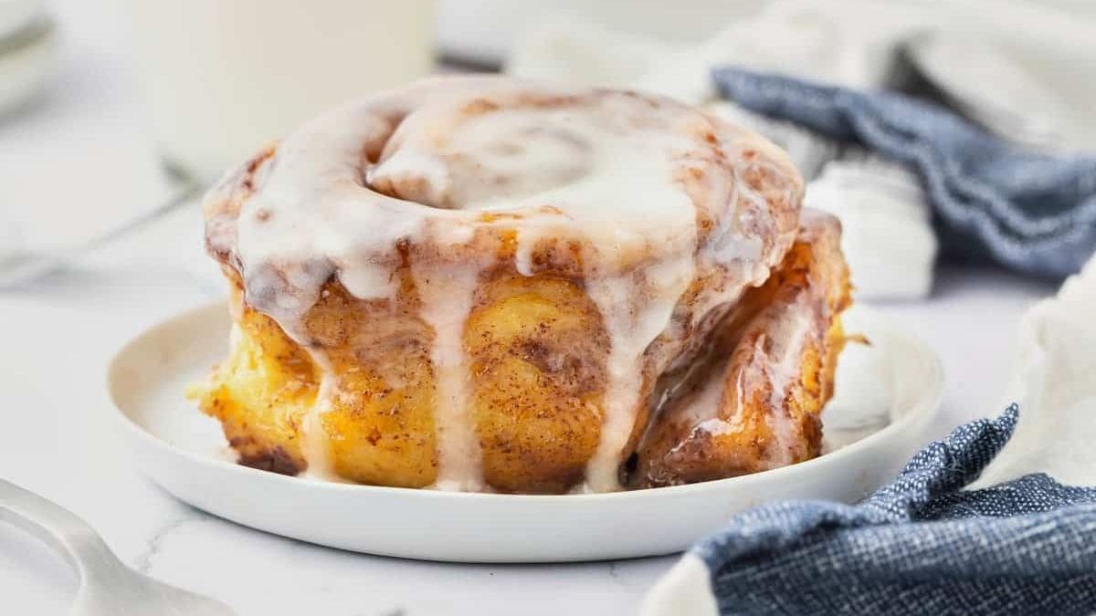 A cinnamon roll covered in white icing sits on a white plate with a blue napkin, all set against a white background.