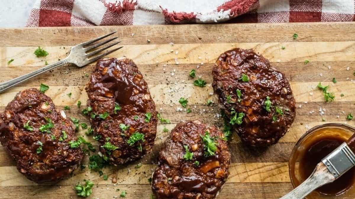 Four Salisbury steaks with brown sauce and chopped parsley are arranged on a wooden cutting board, accompanied by a fork and a small bowl of sauce.