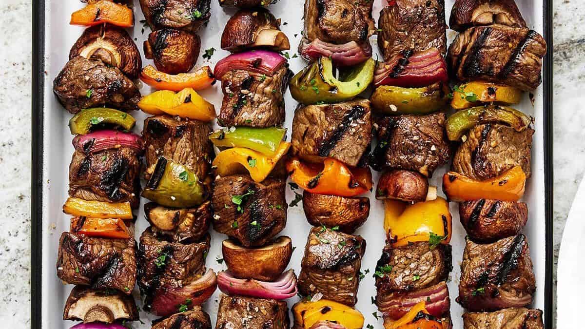Grilled beef kebabs with mushrooms, bell peppers, and red onions arranged in rows on a white tray.
