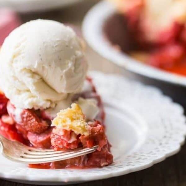 A slice of fruit pie topped with a scoop of vanilla ice cream sits on a white plate with a fork.