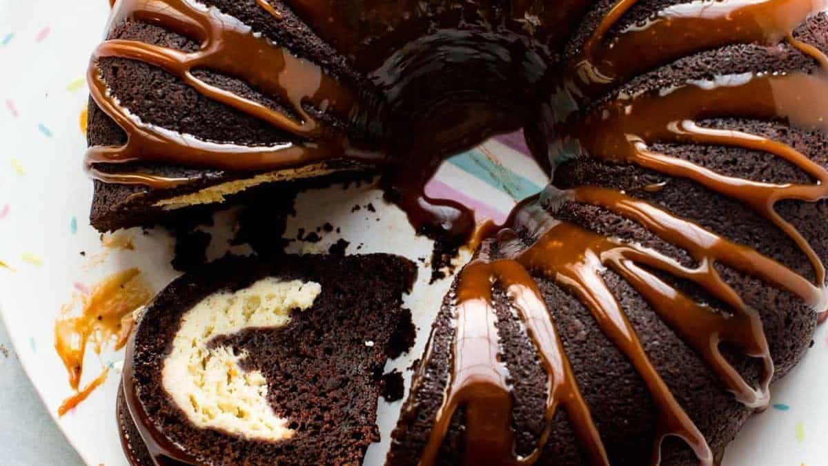 A chocolate bundt cake with a cream cheese swirl and caramel drizzle sits on a plate, with one slice cut and separated from the cake.