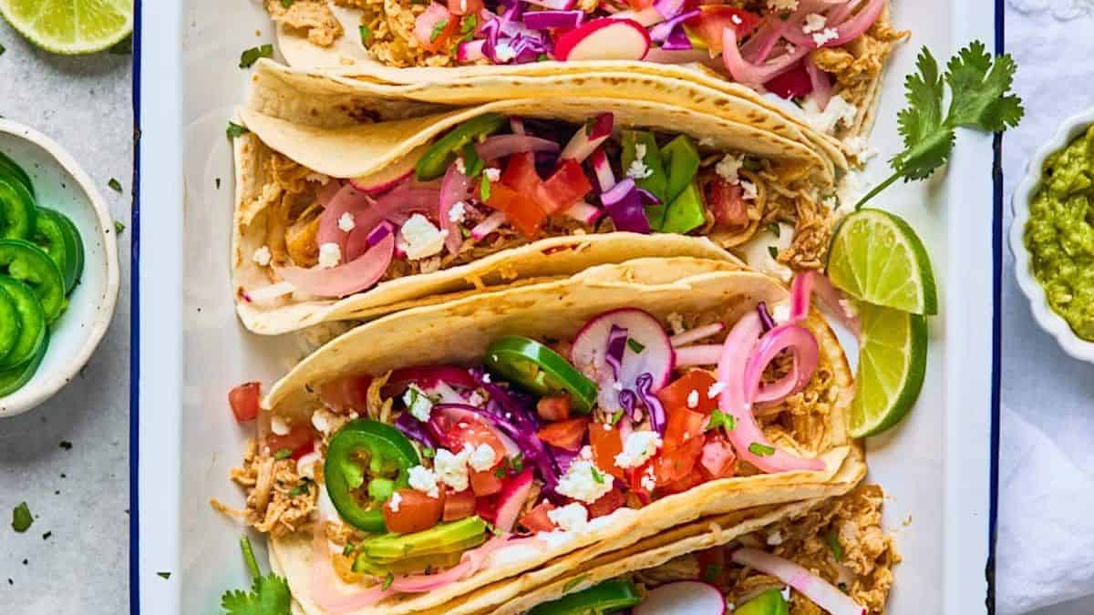 Three soft tacos filled with shredded chicken, sliced jalapeños, radishes, pickled onions, tomatoes, cabbage, and cheese on a rectangular plate. Garnished with lime wedges and cilantro.