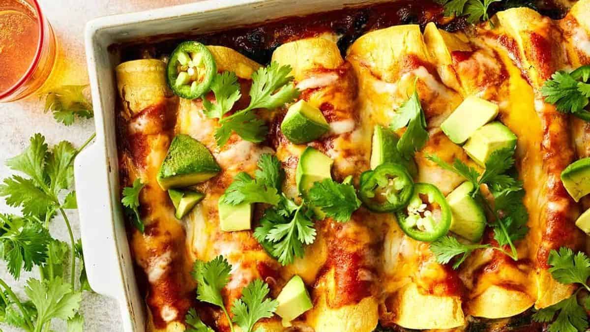 A baking dish filled with enchiladas topped with melted cheese, sliced avocado, jalapeño rounds, and fresh cilantro. A glass of drink and a small bunch of cilantro are placed beside the dish.