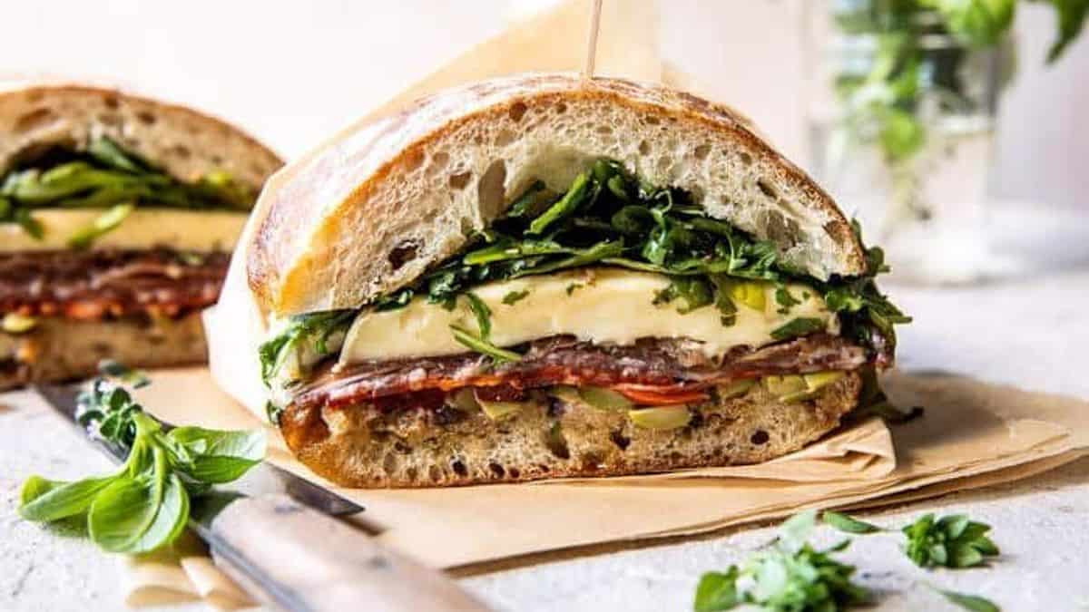 A sandwich with visible layers of cheese, salami, leafy greens, and sliced vegetables on thick rustic bread sits on parchment paper. Beside the sandwich are a knife and sprigs of fresh herbs.