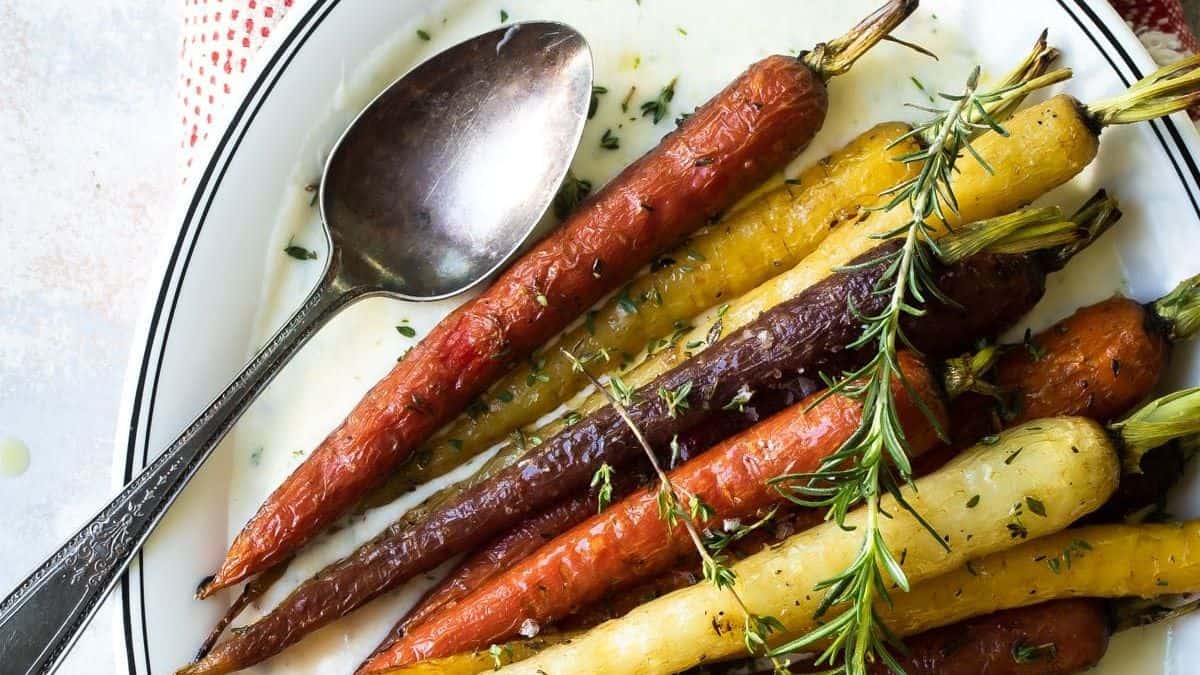 A plate of roasted multicolored carrots garnished with rosemary and herbs, served with a large spoon.