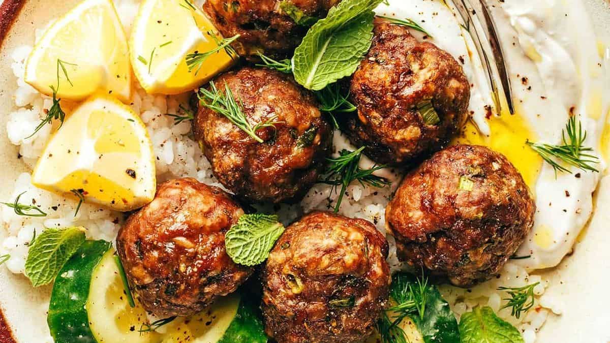 A plate with six meatballs on a bed of rice, garnished with fresh herbs, accompanied by lemon wedges, cucumber slices, and a dollop of yogurt.
