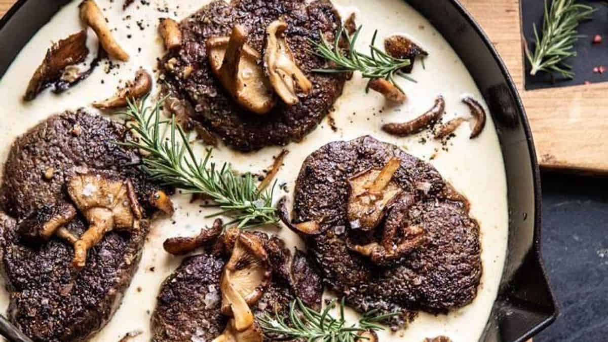 Four seared steaks in a black skillet, topped with mushrooms, sprigs of rosemary, and creamy white sauce.