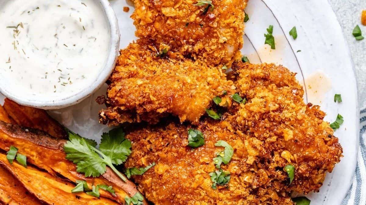A plate with crispy fried chicken, sweet potato fries, fresh cilantro garnish, and a cup of dipping sauce.