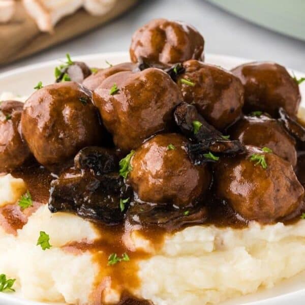A plate of mashed potatoes topped with meatballs, brown gravy, mushrooms, and a garnish of chopped parsley.