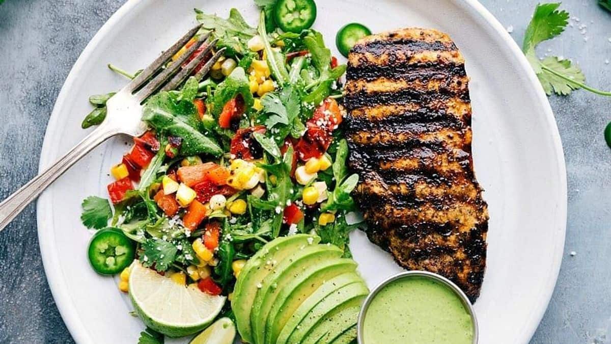 A plate with grilled chicken breast, sliced avocado, mixed salad containing corn and peppers, lime wedges, jalapeño slices, and a small cup of green sauce. A fork rests on the plate.