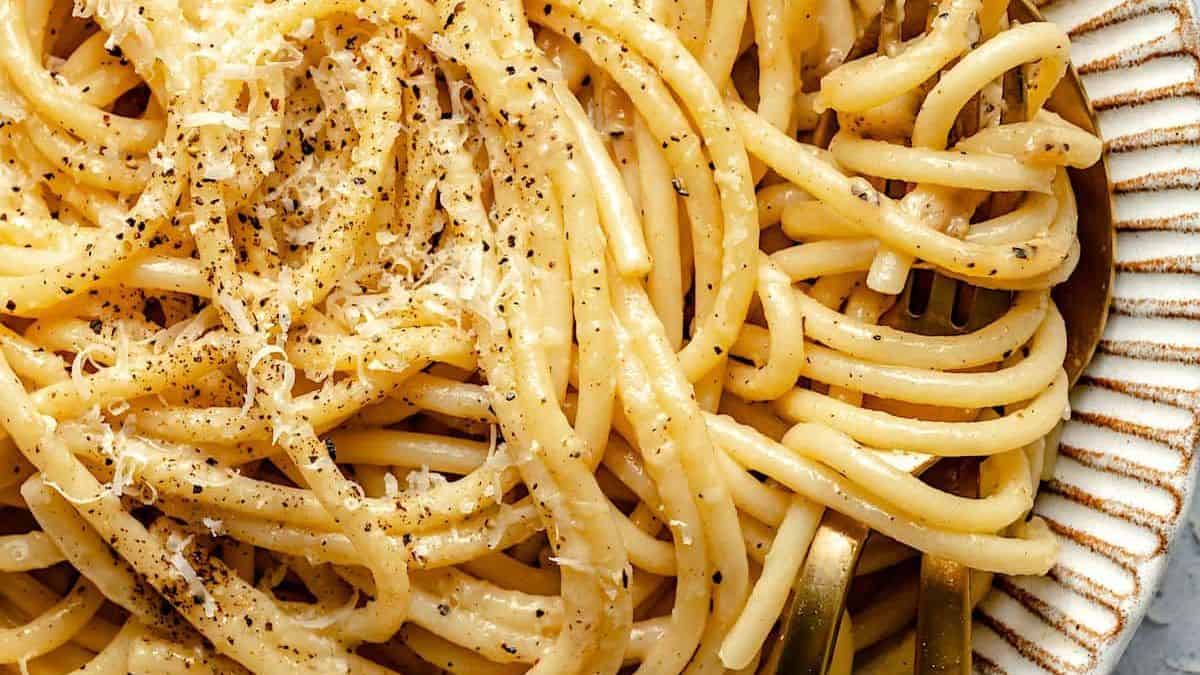 A white plate with cooked pasta, grated cheese, and cracked black pepper, accompanied by gold utensils.