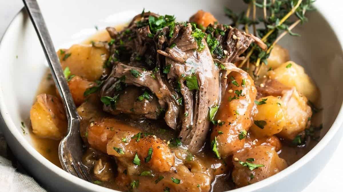 A bowl containing shredded beef, potatoes, carrots, and gravy, garnished with chopped parsley and a sprig of thyme; a spoon rests in the bowl.
