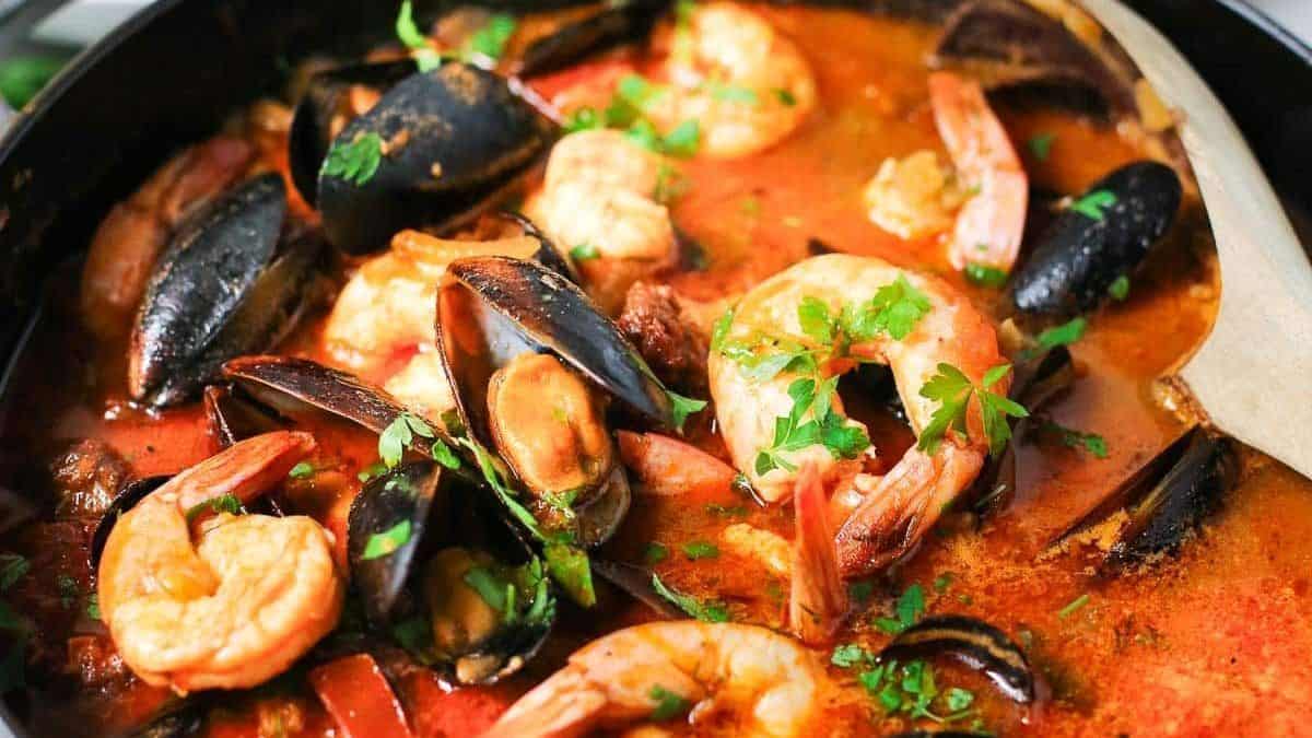 A pan filled with shrimp, mussels, and clams in a red broth, garnished with parsley and accompanied by a wooden spoon.