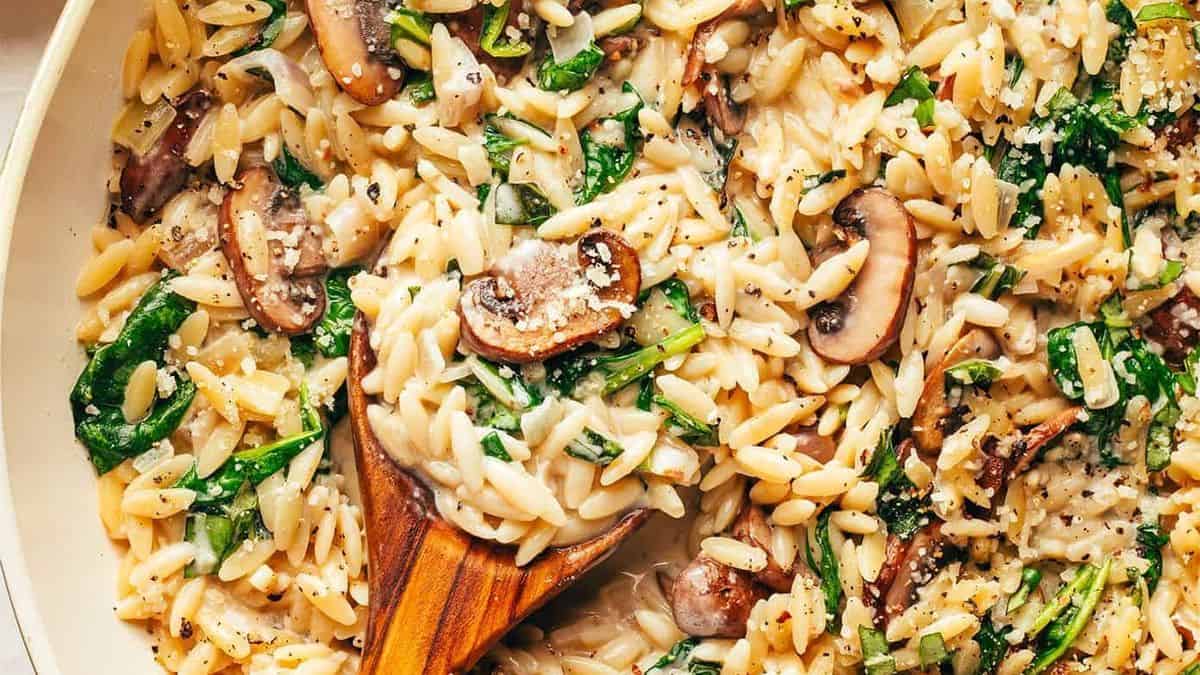Cooked orzo pasta mixed with sautéed mushrooms and spinach, topped with sprinkled cheese, served in a skillet.