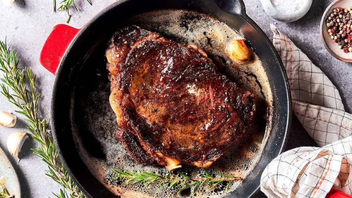 A cooked steak in a cast iron skillet with melted butter, garlic cloves, and rosemary. A checkered napkin and small bowls of peppercorns and salt are beside the skillet.