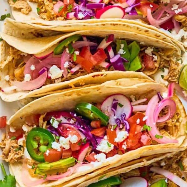 Three chicken tacos filled with shredded chicken, sliced jalapeños, pickled onions, radishes, tomatoes, crumbled cheese, and cilantro, served with lime wedges.