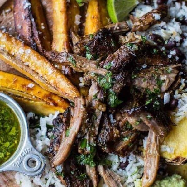 A plate with shredded grilled meat over rice, black beans, fried plantains, pineapple slices, lime wedges, blood orange slices, guacamole, and a small cup of green sauce.