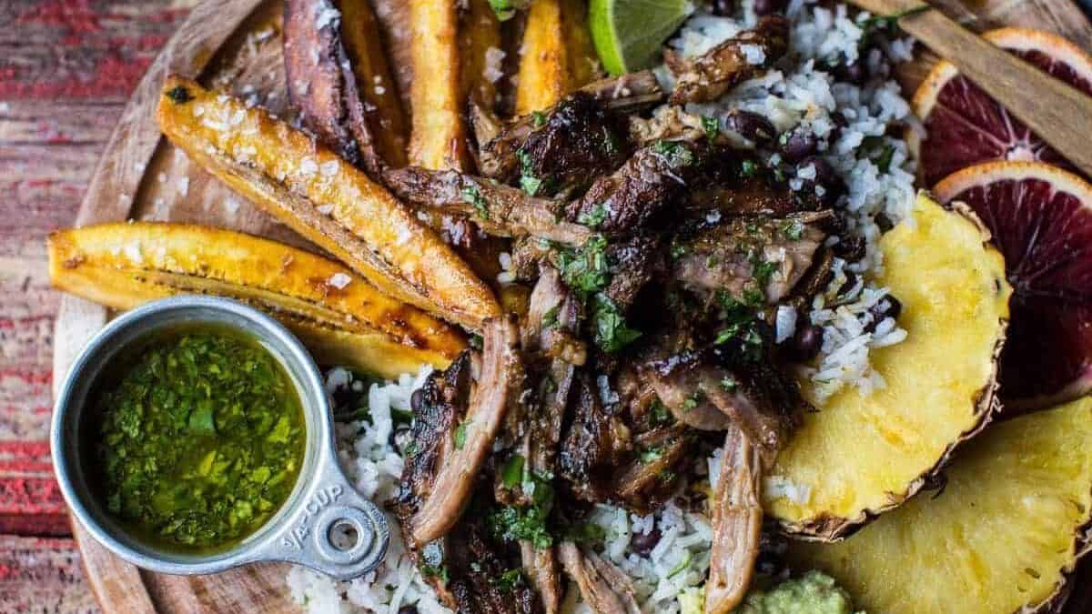 A plate with shredded grilled meat over rice, black beans, fried plantains, pineapple slices, lime wedges, blood orange slices, guacamole, and a small cup of green sauce.