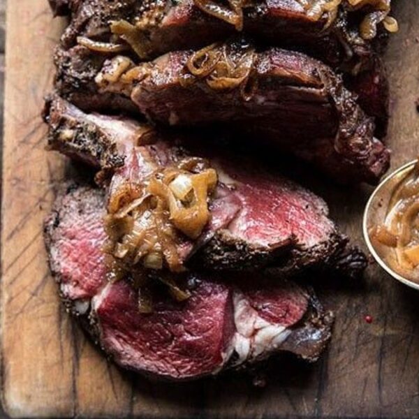 Sliced roast beef topped with caramelized onions on a wooden cutting board, alongside a spoonful of onions and a glass of red drink.