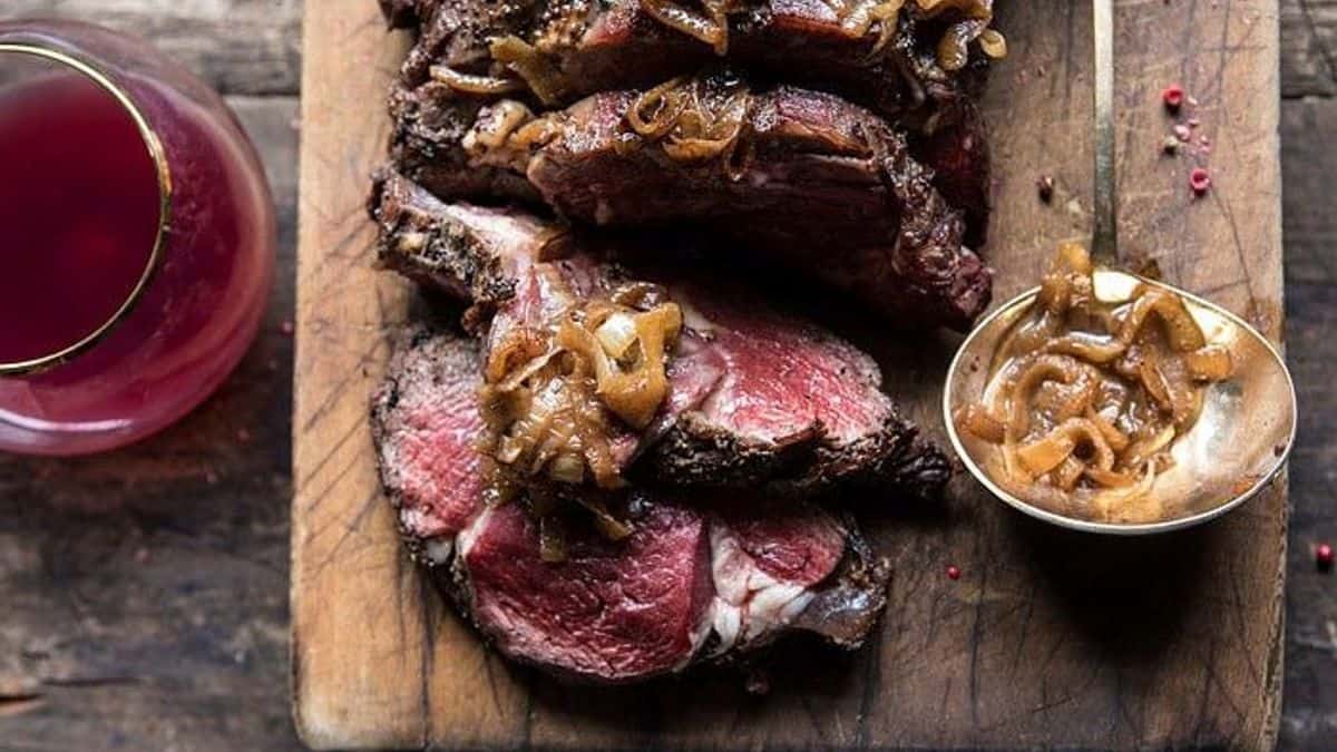 Sliced roast beef topped with caramelized onions on a wooden cutting board, alongside a spoonful of onions and a glass of red drink.