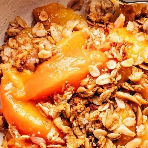 A bowl of peach crisp topped with golden baked oats, visible pieces of peaches and oat crumble, with a spoon.