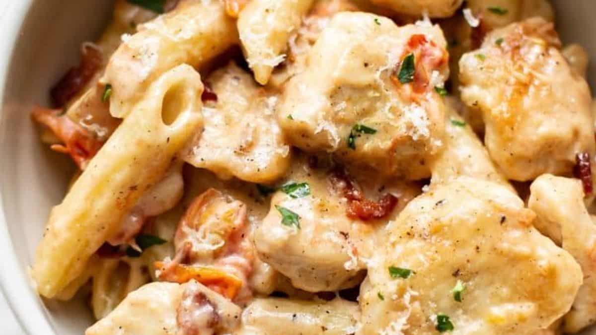 Close-up of creamy penne pasta mixed with chunks of chicken, sun-dried tomatoes, and herbs. The dish is topped with grated cheese and a sprinkle of black pepper.