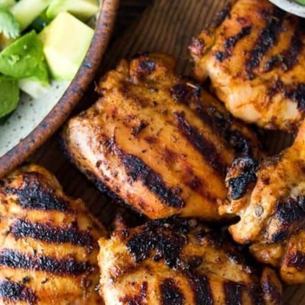 Grilled chicken thighs with char marks rest on a wooden board beside a bowl of salad containing avocado slices, cherry tomatoes, cucumber pieces, and cilantro leaves.
