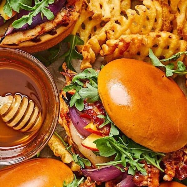 Chicken sandwiches topped with arugula and red onion on brioche buns, accompanied by waffle fries and a bowl of honey with a honey dipper.