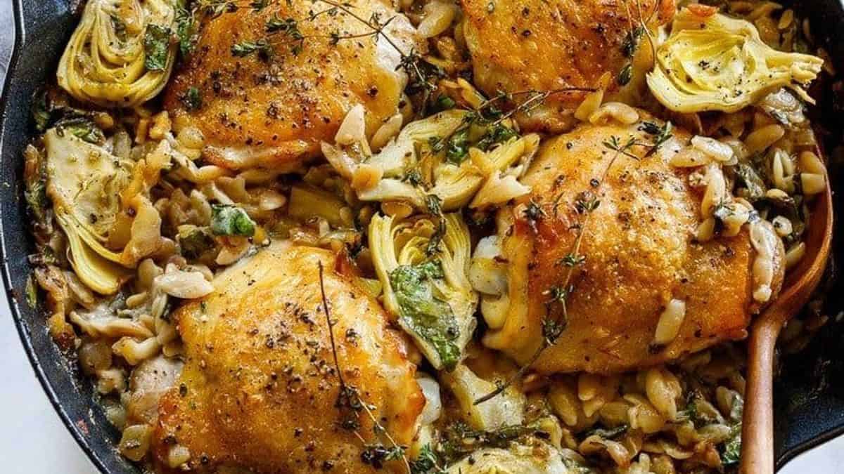 Golden-brown chicken thighs, artichoke hearts, pasta, and herbs in a creamy sauce served in a skillet.