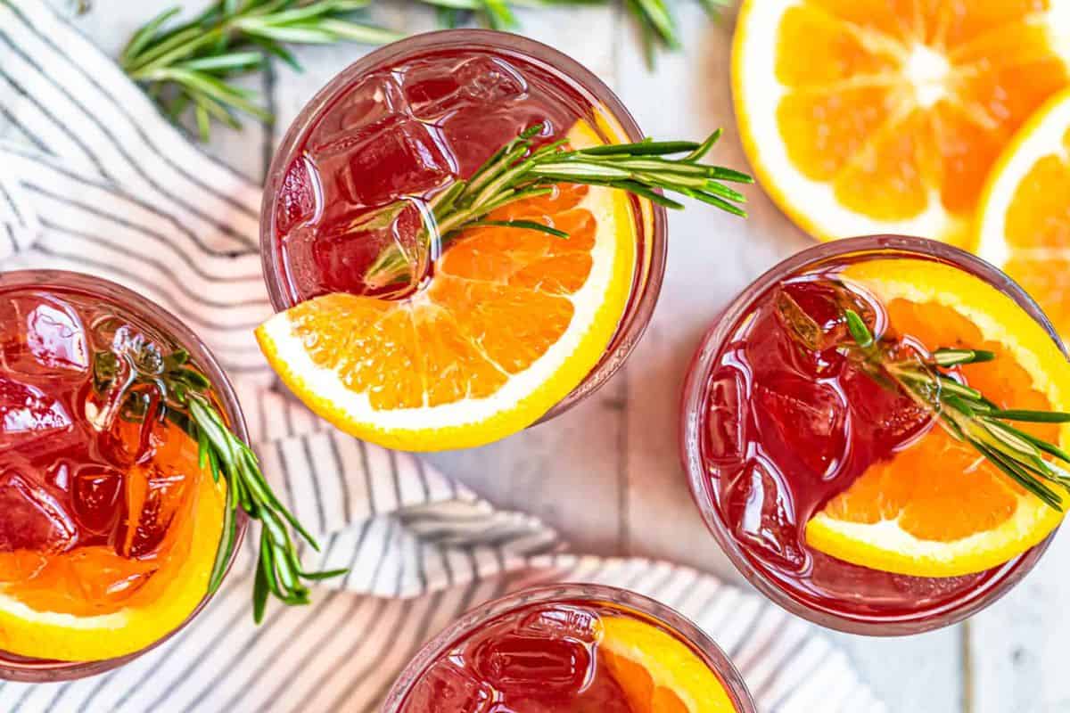 Four glasses of iced red drinks are garnished with orange slices and sprigs of rosemary. They are set on a striped cloth, with extra orange slices nearby.