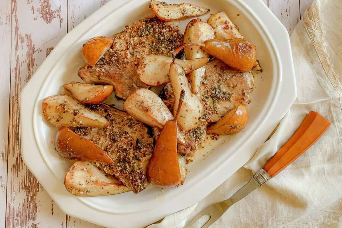 A white serving dish containing baked pork chops and roasted pears, garnished with herbs and spices. The dish is set on a light wood surface next to a knife and fork.