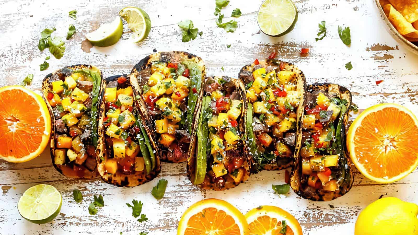 Five grilled tacos topped with diced vegetables and herbs are arranged on a rustic wooden surface. Lime and orange halves, fresh cilantro, and tortilla chips surround the tacos.