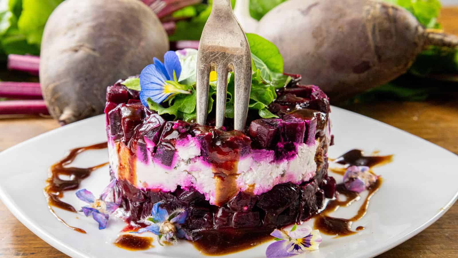 A salad composed of alternating layers of beets and goat cheese, topped with edible flowers and greens, drizzled with balsamic glaze. A fork is inserted on top of the salad, and whole fresh beets are placed in the background.