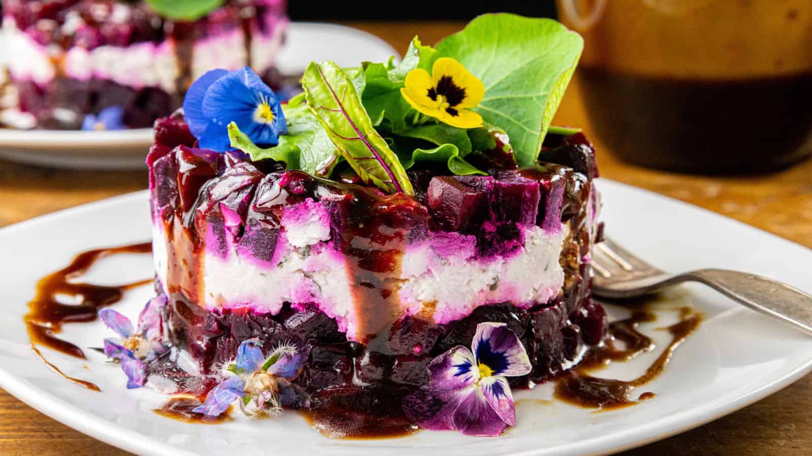 A layered salad with slices of beet and cheese, topped with edible flowers and assorted greens, drizzled with a dark dressing. Presented on a white plate next to a fork.