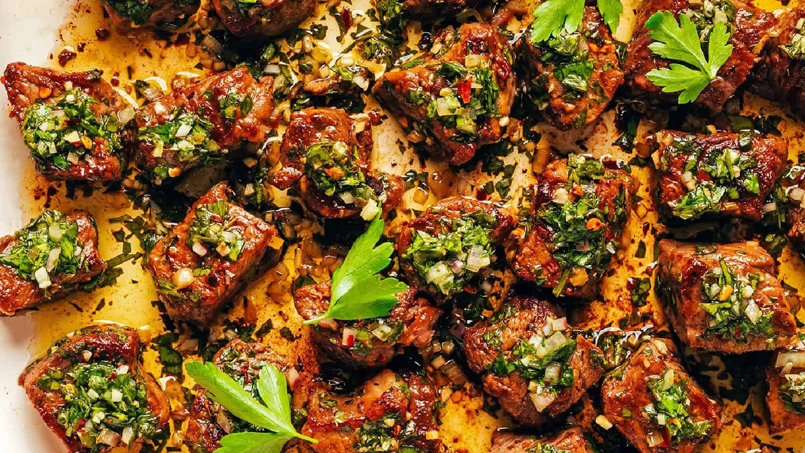 Cooked steak bites topped with chimichurri sauce and garnished with fresh parsley, served on a white platter.