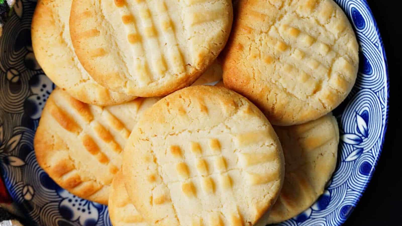 Round, golden-brown cookies with a crisscross fork pattern on top, arranged on a decorative blue and white plate.