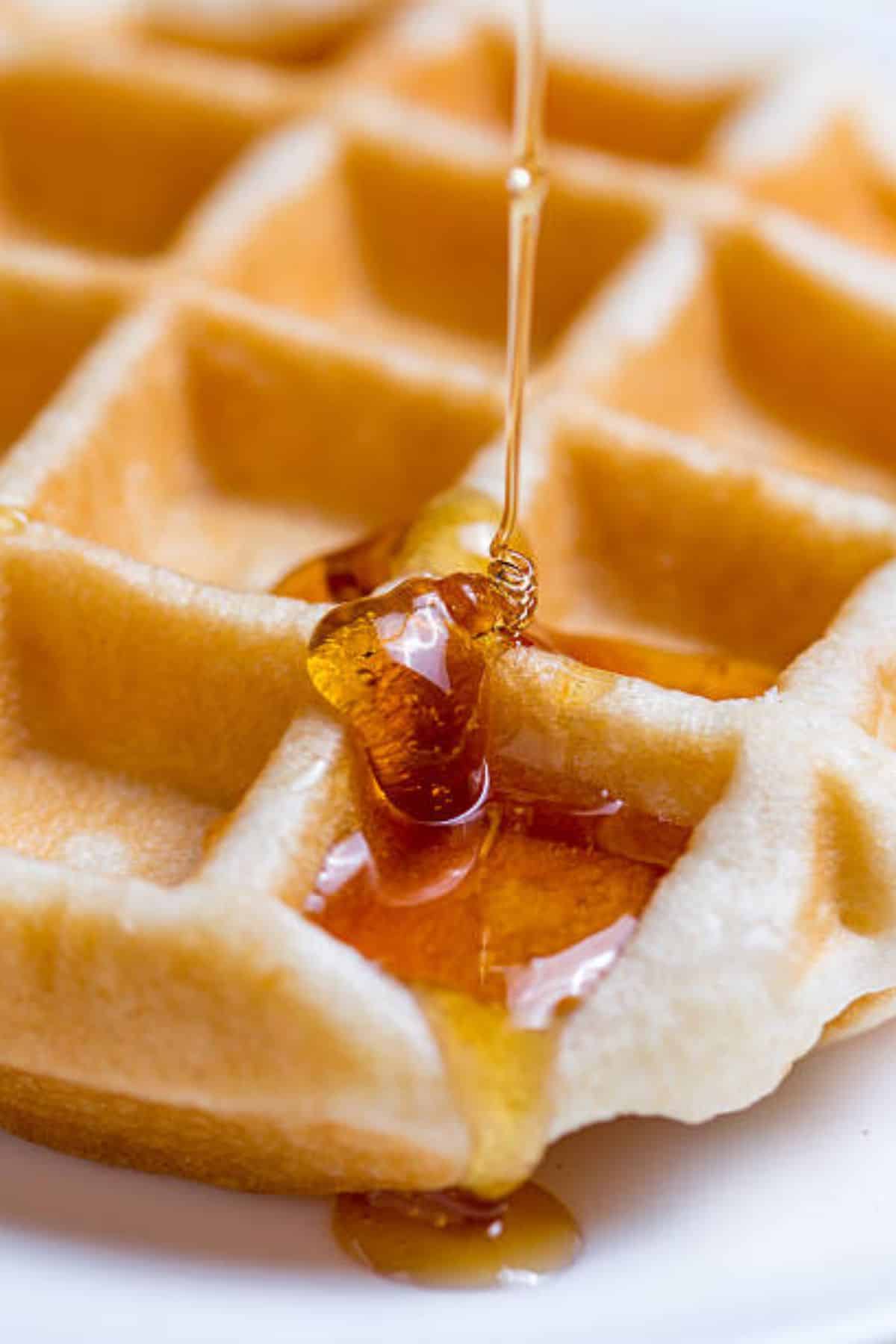 A close-up of maple syrup being poured onto a golden waffle. The syrup pools in the waffle’s square pockets.