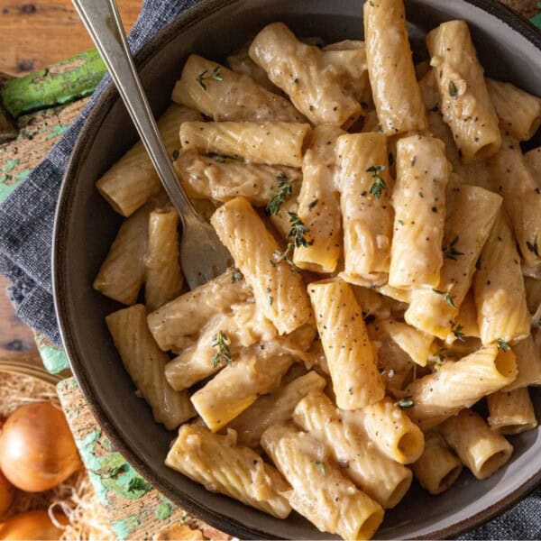 A bowl of rigatoni pasta with creamy sauce, topped with herbs, sits on a wooden table. A fork rests in the bowl. Nearby are piles of shredded cheese and whole onions.